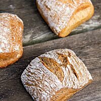 Drei Weizenvollkornbrötchen aus verschiedenen Weizensorten liegen auf einem rustikalen Holztisch.