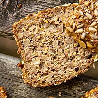 Ein aufgeschnittenes Low-Carb-Brötchen lässt in seiner Krume Kürbis- und Sonnenblumenkerne sowie geschrotete Leinsaat erkennen.
