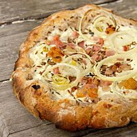 Der mit Schmand, Zwiebeln und Speck belegte Hitzplootz liegt fertig ausgebacken auf einem rustikalen Holztisch.