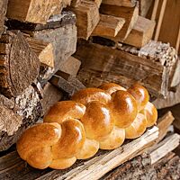 Der goldgelb ausgebackene, glänzende Sauerteigzopf liegt  neben einem Stapel Holzscheite.