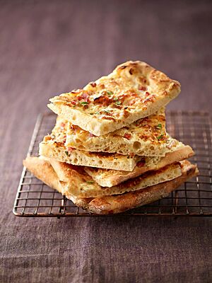 Mehrere Stücke Flammkuchen liegen gestapelt auf einem Gitter, sodass der verhältnismäßig dicke, fluffige Boden gut sichtbar ist.