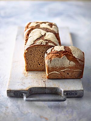 Auf einem Holzbrett liegt das in der Kastenform gebackene Roggenbrot mit weiß bemehlter, rustikaler Kruste und zeigt im Anschnitt seine dunkle, kleinporige Krume.