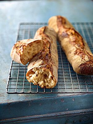Zwei goldbraun ausgebackene und der Länge nach mittig aufgerissene Baguettes mit grobporiger, heller Krume liegen auf einem Rost.