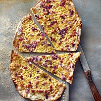 Der dünne und knusprige Roggenflammkuchen ist mit geriebenem Käse, roten Zwiebeln und Speckwürfeln belegt.