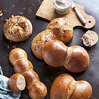 Paarweise aneinander- und goldbraun ausgebacken liegt ein Kölner Röggelchen mit glänzender, glatter Kruste (rechts unten) neben einem Mainzer Wasserweck (rechts mittig), Thüringer Rücklingen (rechts oben) sowie einem Zwiebelbrötchen (links oben) und Münchener Pfenningmuckerln (links unten) auf einem braunen Holztisch.
