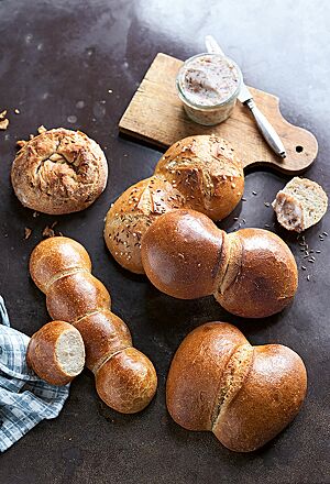 Fünf aneinandergebackene Münchener Pfennigmuckerln mit glatter, glänzender Oberfläche (links unten) liegen neben einem Zwiebelbrötchen (links oben) sowie einem Mainzer Wasserweck (rechts mittig), Thüringer Rücklingen (rechts oben) und Kölner Röggelchen (rechts unten) auf einem braunen Holztisch.
