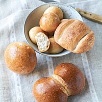Eine Schüssel mit mehreren goldgelb ausgebackenen kleinen Suppenbrötchen (oben) und einem Dresdener Dreierbrötchen (rechts) steht neben einem Kasseler Rundstück (links) und einer Dresdener Franzsemmel (unten) auf einem Tischtuch.