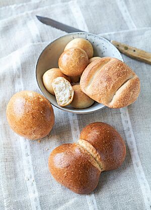 Eine Schüssel mit mehreren goldgelb ausgebackenen kleinen Suppenbrötchen (oben) und einem Dresdener Dreierbrötchen (rechts) steht neben einem Kasseler Rundstück (links) und einer Dresdener Franzsemmel (unten) auf einem Tischtuch.