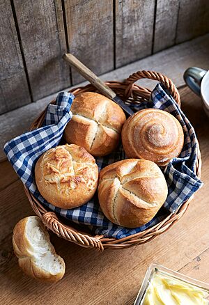 Die runde, goldgelb ausgebackene Münchener Mundsemmel mit kreuzförmigem Ausbund (links oben) liegt neben einem Joghurtbrötchen (rechts oben) sowie einer Rosensemmel (links unten) und einer Kaisersemmel (rechts unten) in einem geflochtenen Brotkorb..
