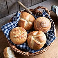 Die runde, goldgelb ausgebackene Rosensemmel mit rustikal aufgerissener Kruste (links unten) liegt neben einer Kaisersemmel (rechts unten) sowie einer Münchener Mundsemmel (links oben) und einem Joghurtbrötchen (rechts oben) in einem geflochtenen Brotkorb.