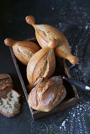 Ein länglich geformtes Baguettebrötchen mit rustikal aufgerissener, bemehlte Kruste und grober, unregelmäßige Porung (unten) liegt mit drei Fränkischen Kipfen (oben) in einer eckigen Holzschale.