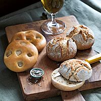 Zwei goldgelb ausgebackene Speckwecken mit auf der glatten Oberfläche sichtbaren Speckwürfeln (links) liegen neben drei Maurerlaibln (rechts) und einem Glas Bier (oben) auf einem Holzbrett.
