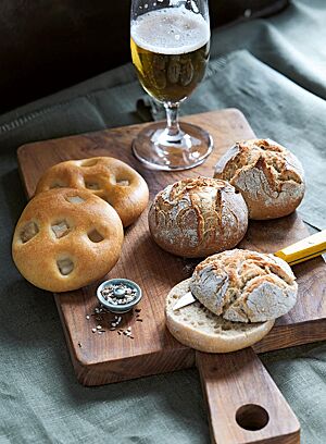 Zwei goldgelb ausgebackene Speckwecken mit auf der glatten Oberfläche sichtbaren Speckwürfeln (links) liegen neben drei Maurerlaibln (rechts) und einem Glas Bier (oben) auf einem Holzbrett.