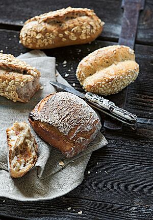 Ein längliches, rustikal aufgerissenes Müslibrötchen mit Haferflocken auf der Kruste (oben) liegt neben einem Bergsteigerbrötchen (unten) und einem Haferbollen (Mitte) auf einem Leinentuch.