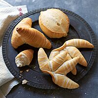 Zwei goldgelb ausgebackene Fingerbrötchen mit gewickelter Struktur und glänzender, glatter Oberfläche (rechts) liegen mit einem Mailänder Brötchen (Mitte) und zwei Cornetti (links) auf einer dunklen Platte.