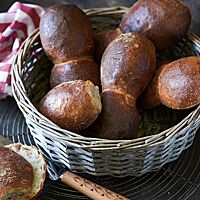 In einem hellgrauen Brotkorb liegen mehrere paarweise angebackene Bürli. Die Kruste ist stark glänzend und dunkel ausgebacken. Ein aufgeschnittenes Bürli liegt auf einem Gitterrost vor dem Brotkorb und zeigt die luftig-lockere, helle Krume.