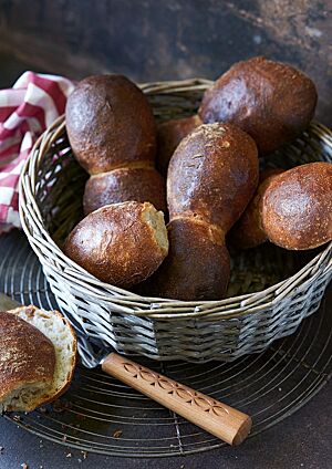 In einem hellgrauen Brotkorb liegen mehrere paarweise angebackene Bürli. Die Kruste ist stark glänzend und dunkel ausgebacken. Ein aufgeschnittenes Bürli liegt auf einem Gitterrost vor dem Brotkorb und zeigt die luftig-lockere, helle Krume.