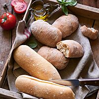 Ein grau-braunes Leinentuch mit vier Ciabattini und zwei Panini liegt auf einem Holztablett. Die Ciabattini (oben) haben eine glatte, leicht bemehlte Kruste und eine luftig-lockere Krume mit einer mittleren bis groben Porung.