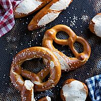 Auf einem dunklen Backblech mit Lochung liegen Laugenbrezeln (Mitte), Laugenstangen (oben) und Laugenbrötchen (unten). Ihre glänzende, feine Kruste ist dunkelbraun ausgebacken und mit grobem Salz bestreut.