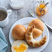 Auf einem weißen Frühstücksteller liegen zwei Milchbrötchen mit glatter (oben links) und gedrückter Oberseite (oben rechts), eine mit Marmelade bestrichene Milchbrötchenhälfte (unten links) und ein Milchhörnchen (unten rechts). Das gedrückte Milchbrötchen hat eine goldgelb ausgebackene, leicht glänzende Oberfläche, die mittig zweigeteilt ist.