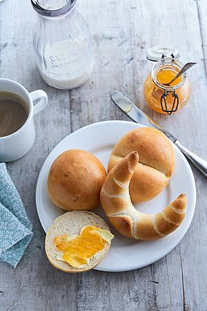 Auf einem weißen Frühstücksteller liegen zwei Milchbrötchen mit glatter (oben links) und gedrückter Oberseite (oben rechts), eine mit Marmelade bestrichene Milchbrötchenhälfte (unten links) und ein Milchhörnchen (unten rechts). Das Milchhörnchen hat eine goldgelb ausgebackene, leicht glänzende Oberfläche, auf der die Windungen durch die Wicklung beim Formen zu sehen sind.