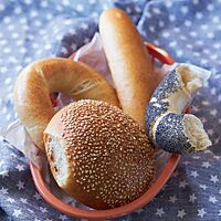 Auf einer blauen Decke mit weißen Sternen steht ein Brotkorb, der mit einem Sandwich-Brötchen (oben), eineinhalb Bagels (links und rechts) und einem Hamburger Bun (unten) gefüllt ist. Der Bagel hat eine stark glänzende, goldgelb ausgebackene, feine Kruste und eine lockere, weiche Krume.