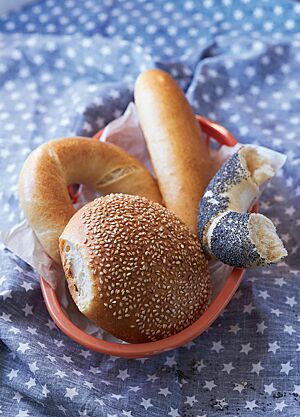 Auf einer blauen Decke mit weißen Sternen steht ein Brotkorb, der mit einem Sandwich-Brötchen (oben), eineinhalb Bagels (links und rechts) und einem Hamburger Bun (unten) gefüllt ist. Der Bagel hat eine stark glänzende, goldgelb ausgebackene, feine Kruste und eine lockere, weiche Krume.