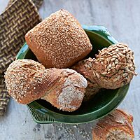 In einer grünen Schüssel liegen ein Weizenvollkornbrötchen (Mitte) und  zwei Saatenschrotlinge (rechts und links). Die in sich verdrehten, länglichen Saatenschrotlinge haben eine in Schrot bzw. Sonnenblumenkernen gewälzte Oberfläche.