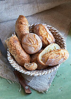 Ein mit Dinkelbrötchen (Mitte), Dinkelkartoffelbrötchen (rechts) und Dinkelseelen (links) gefüllter Brotkorb steht auf einem braunen Tuch. Die länglichen Dinkelkartoffelbrötchen haben eine rustikal aufgerissene, leicht bemehlte, krosse Kruste und eine mittelporige, elastische Krume.