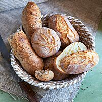 Ein mit Dinkelbrötchen (Mitte), Dinkelkartoffelbrötchen (rechts) und Dinkelseelen (links) gefüllter Brotkorb steht auf einem braunen Tuch. Die langen Dinkelseelen haben eine glatte, glänzende, mit Kümmel und grobem Salz bestreute Kruste.