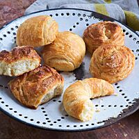 Auf einer weißen Emailleplatte liegen Splitterhörnchen (unten rechts), Dänische Brötchen (oben links) und Splitterbrötchen in der Ost- und Westvariante (unten links und oben rechts). Die leicht eckigen Dänischen Brötchen haben eine goldgelb ausgebackene, glänzende Kruste.