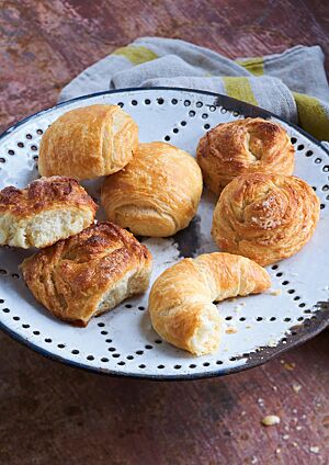 Auf einer weißen Emailleplatte liegen Splitterhörnchen (unten rechts), Dänische Brötchen (oben links) und Splitterbrötchen in der Ost- und Westvariante (unten links und oben rechts). Die leicht eckigen Dänischen Brötchen haben eine goldgelb ausgebackene, glänzende Kruste.