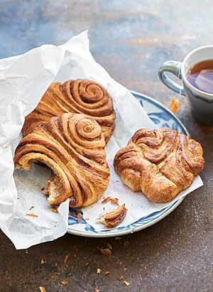 Auf einem blau-weißen Teller liegen zwei gerollte Franzbrötchen (links) und ein gedrücktes Franzbrötchen (rechts) in einer Frühstückstüte. Das gerollte Franzbrötchen hat eine zart-knusprige Kruste und eine saftige Krume.
