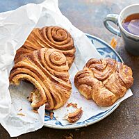 Auf einem blau-weißen Teller liegen zwei gerollte Franzbrötchen (links) und ein gedrücktes Franzbrötchen (rechts) in einer Frühstückstüte. Das gedrückte Franzbrötchen mit wilder, zufälliger Form hat eine zart-knusprige Kruste und eine saftige Krume.