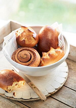 In einer weißen, mit einem Tuch ausgelegten Schüssel liegen zwei Eiwecken (oben links und unten rechts), ein Rosinenbrötchen (oben rechts) und ein Zimtweck (unten links). Die Eiwecken haben eine goldgelb bis goldbraun ausgebackene, glänzende Kruste mit einem Zackenmuster auf der Oberfläche.