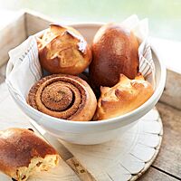 In einer weißen, mit einem Tuch ausgelegten Schüssel liegen zwei Eiwecken (oben links und unten rechts), ein Rosinenbrötchen (oben rechts) und ein Zimtweck (unten links). Das längliche Rosinenbrötchen hat eine glänzende, dunkelbraun ausgebackene, feine Kruste.