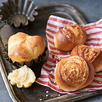 Auf einem rot-weiß-gestreiften Papier liegen drei Campingbrötchen (rechts) und eine Brioche (links) auf einem Tablett. Die Campingbrötchen haben eine goldbraun ausgebackene, glänzende Kruste mit Hagelzucker, der im weiten Ausbund an die Oberfläche kommt.
