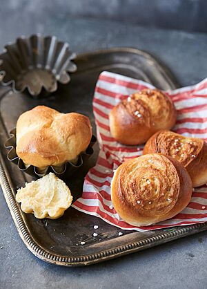 Auf einem rot-weiß-gestreiften Papier liegen drei Campingbrötchen (rechts) und eine Brioche (links) auf einem Tablett. Die Campingbrötchen haben eine goldbraun ausgebackene, glänzende Kruste mit Hagelzucker, der im weiten Ausbund an die Oberfläche kommt.