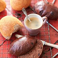 Auf einer roten Arbeitsfläche liegt ein rechteckiger Gitterrost mit Schokobrötchen im Ganzen (oben rechts) und im Anschnitt (unten) sowie Kaneelbrötchen (oben links) und einer Tasse Kaffee. Die länglich geformten, glänzenden Schokobrötchen haben eine glatte, dünne Kruste und eine kleinporige, dunkle Krume.