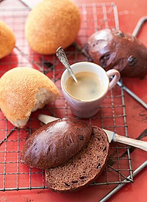 Auf einer roten Arbeitsfläche liegt ein rechteckiger Gitterrost mit Schokobrötchen im Ganzen (oben rechts) und im Anschnitt (unten) sowie Kaneelbrötchen (oben links) und einer Tasse Kaffee. Die in Zimt, Zucker und Semmelbröseln gewälzten Kaneelbrötchen haben eine goldgelb ausgebackene Kruste und eine helle, wattige Krume.