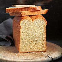 Zwei Scheiben Butterstuten liegen auf dem angeschnittenen, goldbraun ausgebackenen Stuten, der auf einem Holzbrett liegt. Die Krume des Stutens ist wattig und elastisch.