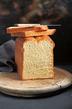 Zwei Scheiben Butterstuten liegen auf dem angeschnittenen, goldbraun ausgebackenen Stuten, der auf einem Holzbrett liegt. Die Krume des Stutens ist wattig und elastisch.