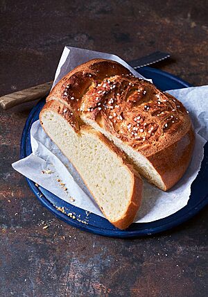 Das angeschnittene Campingbrot liegt auf Pergamentpapier auf einer blauen Platte. Die helle Krume ist wattig-weich, die rustikale Kruste goldbraun ausgebacken. Im Ausbund des Brotes kommt der eingearbeitete Hagelzucker zum Vorschein.