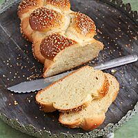 Eine Challah liegt angeschnitten mit zwei Scheiben im Vordergrund auf einem Holztablett mit feiner Verzierung. Die Krume der Challah ist wattig und saftig. Die Windungen des Zopfes sind dunkelbraun ausgebacken und mit Sesam bestreut.
