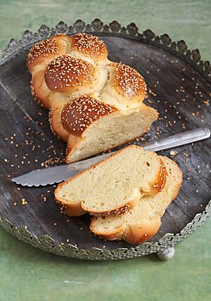 Eine Challah liegt angeschnitten mit zwei Scheiben im Vordergrund auf einem Holztablett mit feiner Verzierung. Die Krume der Challah ist wattig und saftig. Die Windungen des Zopfes sind dunkelbraun ausgebacken und mit Sesam bestreut.