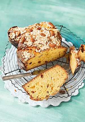 Eine angeschnittene Colomba liegt auf einem Gitterrost. Im Anschnitt wird die wattig-weiche Krume mit Sultaninen und Orangeat sichtbar. Auf der Oberfläche der Colomba ist das Topping aus Eiklar, Zucker und Mandeln erkennbar.