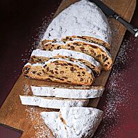 Ein mit reichlich Puderzucker bestäubter, angeschnittener Stollen liegt auf einem langen Holzbrett. Die Scheiben des Stollens zeigen die von Zitronat und Sultaninen durchzogene, saftige Krume.