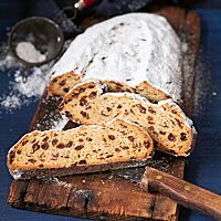 Zwei große Scheiben des Erzgebirgischen Stollen liegen auf einem rustikalen, dunklen Holzbrett. Die Krume des Stollens ist von vielen Sultaninen, Orangeat und Zitronat durchzogen. Die weit aufgerissene Kruste ist kräftig mit Feinkristallzucker bestreut.