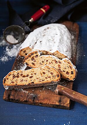 Zwei große Scheiben des Erzgebirgischen Stollen liegen auf einem rustikalen, dunklen Holzbrett. Die Krume des Stollens ist von vielen Sultaninen, Orangeat und Zitronat durchzogen. Die weit aufgerissene Kruste ist kräftig mit Feinkristallzucker bestreut.