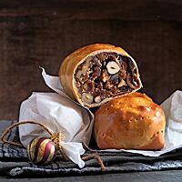 Ein angeschnittenes Früchtebrot mit dunklem Kern und dünnem, hellem Mantel liegt auf einem Früchtebrot im Ganzen und einem in Pergamentpapier eingewickelten Früchtebrot. Im hellen Kern des Früchtebrotes sind Haselnüsse und Walnüsse in der Fruchtmasse erkennbar.